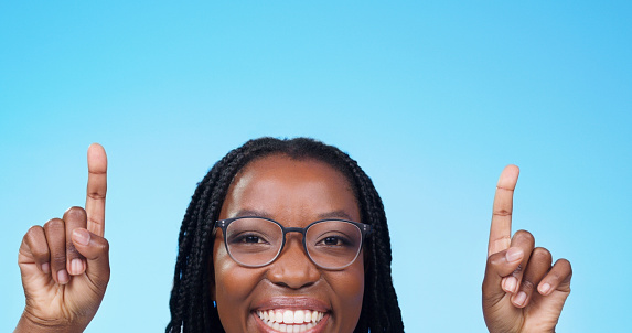 Smile, happy black woman pointing fingers up and in studio background. Closeup female face, African person gesturing at product placement and showing information space or notification on blue screen