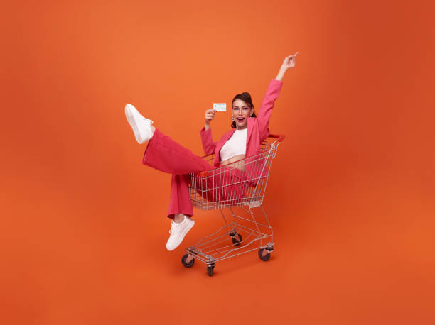 portrait en pied d’une femme à la mode souriante heureuse avec chariot et montrant une carte de crédit isolée sur fond orange - lenght photos et images de collection