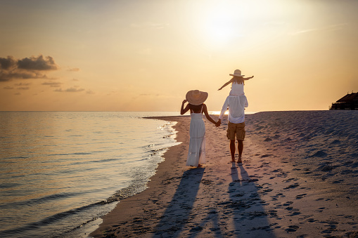 A elegant family in white clothing walks hand in hand down a tropical paradise beach during sunset tme and enjoys their summer vacatios