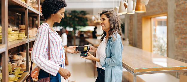 Happy ceramic store owner receiving a contactless NFC payment from a customer in her shop. Successful small business owner scanning a smartphone on a credit card machine while serving a customer.