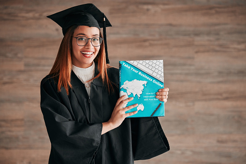 Graduate, woman portrait and business textbook of a young student happy from graduation. Learning book, happiness and excited female ready for university education with a smile from knowledge