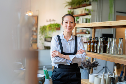 Customer service builds a company's image. Manager owner wearing apron standing in the home decoration shop filled with items and smile during working day with arm crossed. Many ceramic and glass product on the shelf
