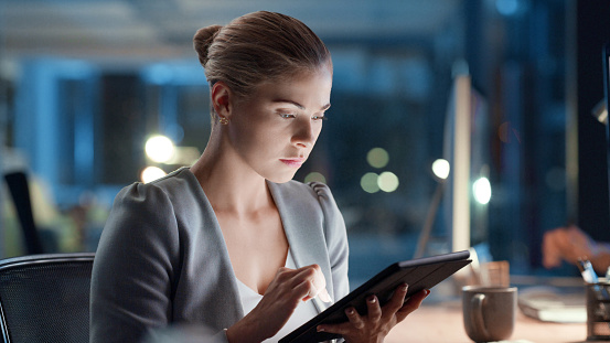 Software analyst browsing a tablet while working on a computer late at night in modern office. Woman doing research, testing an app or communicating with developers. Female focused on an online task