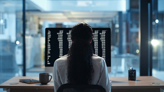 Business woman working through data, looking at stats and meeting a deadline while working late in office alone from behind. Corporate female, professional and analyst doing research online at work