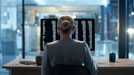 Back view of programmer or cybersecurity employee working overtime late at night in an office. Skilled woman web expert writing a backend algorithm on a computer in the evening at her workplace