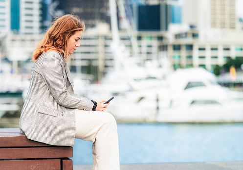 Profile view of a woman as she sits down to use her phone by the marina in Auckland's Viaduct Harbour.