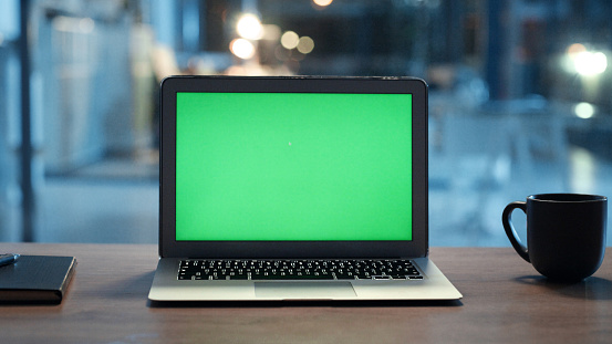 Closeup of chroma key green screen on a laptop or empty CGI, copyspace desktop computer display template. Mock up pc monitor on office desk at night, alongside a note book, pen and cup or mug.