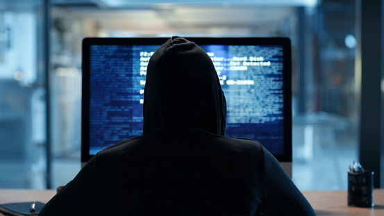 Back of hacker or cyber criminal stealing information online sitting at a computer at night. Hooded mysterious person coding spyware, malware and viruses to commit fraud, theft and phishing scam