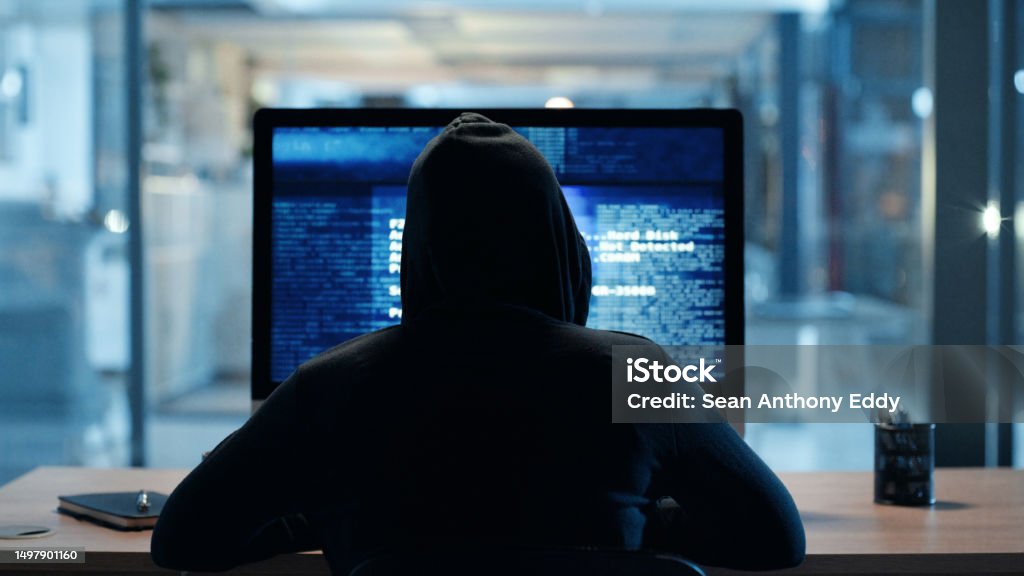 Back of hacker or cyber criminal stealing information online sitting at a computer at night. Hooded mysterious person coding spyware, malware and viruses to commit fraud, theft and phishing scam Computer Hacker Stock Photo