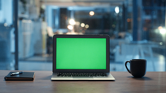 Laptop screen with green, blank chromakey on desk in office late at night. Blurred background of workplace with furniture and equipment. Empty work environment late in the evening