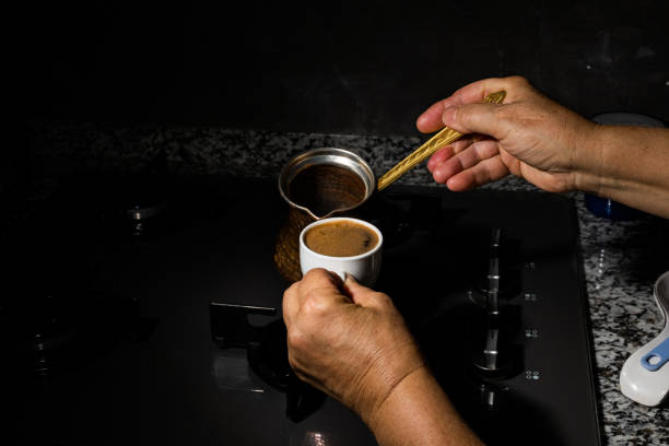 Coffee pot on the stove in the kitchen traditional Turkish coffee is being cooked in a traditional coffee pot, with plenty of foam. Background is dark, space for text.A hand is pouring coffee into a cup.