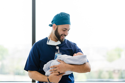After assisting in a difficult delivery, the young adult male surgeon smiles down at the newborn in his arms.