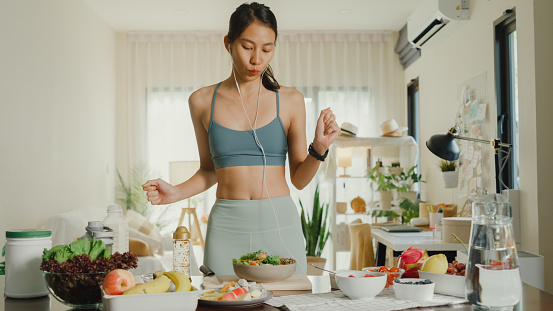 Young Asian athletic woman wearing headset sing stereo volume song listen music and cooking salad at home. Diet and healthy food concept.
