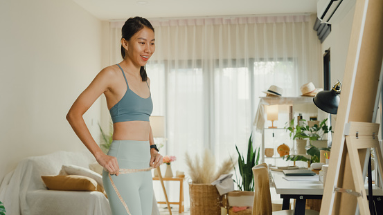 Young Asian athletic woman with a tape measure body perfect slim weight loss in front of a mirror in living room at home. Diet and healthy weight loss concept.