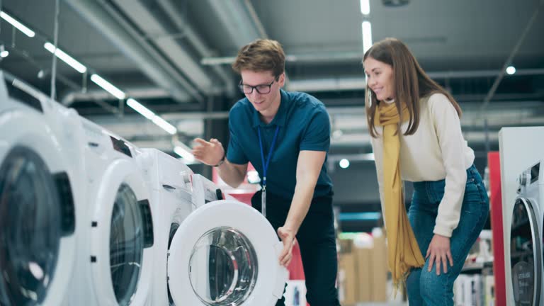 Portrait of a Retail Electronics Shop Consultant Assisting Young Female Customer in Selecting a Washing Machine. Beautiful Female Evaluating Modern Laundry Solutions