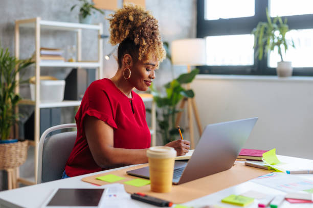 Side profile of young beautiful African-American business woman writing down notes while sitting at office desk. stock photo