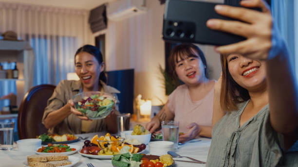 Young Asian women friends eating salad and holding mobile video call online sit at dining table in dining room at home at night. Lifestyle healthy food eating enjoy life and plant-based diet concept. jóvenes amigas asiáticas comiendo ensalada y realizando videollamadas móviles en línea se sientan en la mesa del comedor en el comedor de su casa por la noche. estilo de vida alimentos saludables comer disfrutar de la vida y la dieta basada en plantas.
