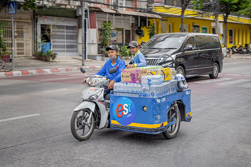 Bangkok, Thailand - March 28th 2023: Two men driving down a road on a motorcycle with a sidecar heavy loaded with waterbottles in the center of the capital of Thailand
