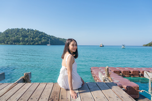 Beautiful young asian woman in white dress sitting on wooden pier among tropical sea in summer vacation at Koh Kood Island