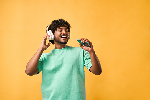 Portrait of a curly Indian man in a turquoise t-shirt singing with a phone in wireless headphones enjoying music.
