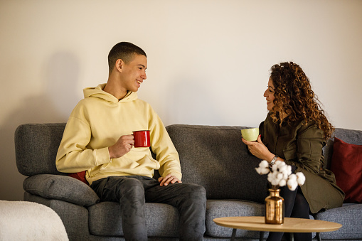 Portrait of happy teenage boy and his mother sitting on the sofa in their living room casually chatting over cups of coffee or tea.