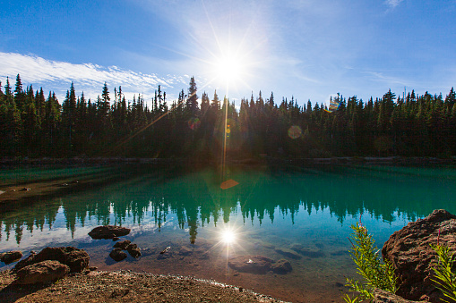 Natures getaway with a sun star reflecting off bright still turquoise lake surrounded by pine trees in Canada.