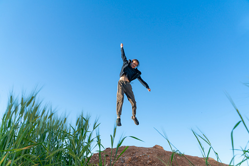 young boy jumping into the air above the hill. Ears of wheat are seen in the foreground. Taken from the bottom angle. Shot with a full-frame camera in daylight.