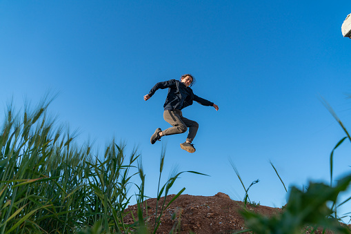 young boy jumping into the air above the hill. Ears of wheat are seen in the foreground. Taken from the bottom angle. Shot with a full-frame camera in daylight.