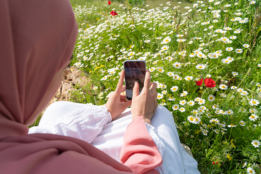 woman in headscarf taking photo with mobile phone among wildflowers in spring. Woman in a white long dress. having a good time among daisy and poppy flowers.Shot with a full-frame camera in daylight.