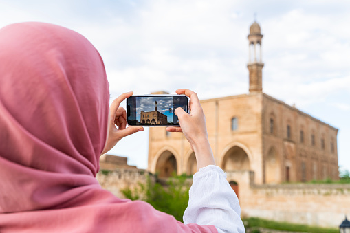 A woman in a headscarf taking a photo with her mobile phone in the view of the historical church in Midyat district of Mardin. woman is sitting at the table on the terrace. Shot with a full-frame camera on a sunny day.