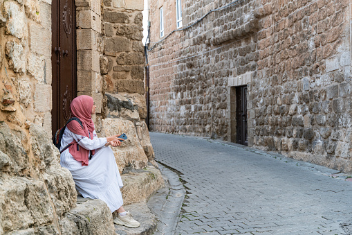 Tourist woman sitting on the steps of the stone paved streets between the stone houses of Midyat district of Mardin province. Tourist woman wearing a headscarf is walking around the historic stone streets. sitting on the stair step at the door entrance using a cell phone. Shot with a full-frame camera in daylight.