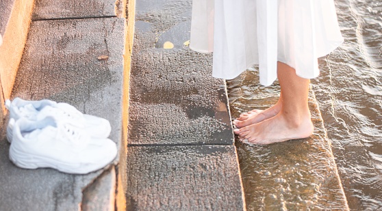 A female wearing a white dress with bare feet near a water and her shoes on the stairs