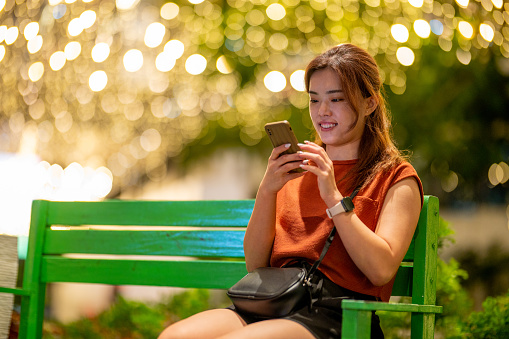 An Asian woman is sitting and relaxing on a park bench in the city, using a smartphone and a digital tablet at evening. Humanizing digital experiences