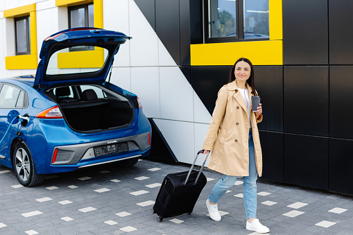 A young girl stopped to charge her electric car, and in the meantime goes to rest, carrying a suitcase.