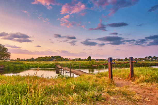 Landscape with sunset over river. Rural pedestrian old bridge. Evening with calm beautiful clouds on sky. Water reflection. Tranquil evening scene. Green filed and meadow. Europe, Belarus