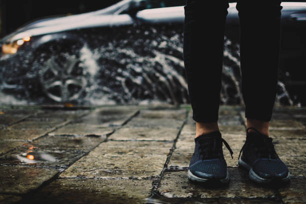 istock Blurred splashes flying all around as grey metallic German car of a modern design drives into a puddle. Girl wearing dark blue sneakers is standing on the sidewalk by the road, sharp reflection of a car and its headlights in a small puddle. 1497966277