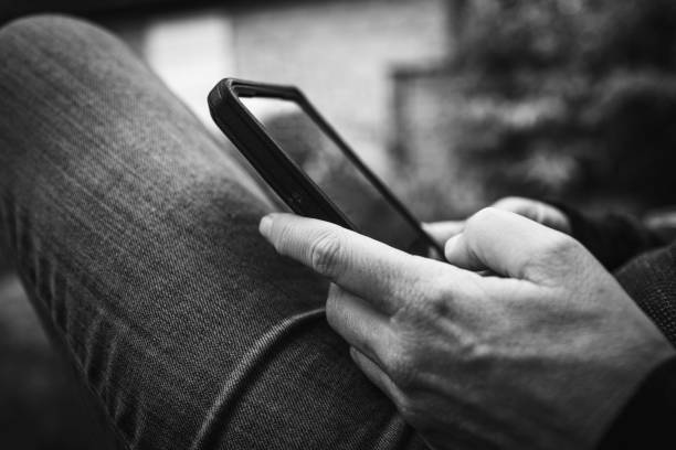 gros plan d’une personne vérifiant un smartphone assise sur un banc extérieur dans un jean décontracté. voyager, se connecter, se détendre, travailler, en ligne, e-work, remort work concepts. photo rétro noir et blanc. - retro revival telephone human hand toned image photos et images de collection