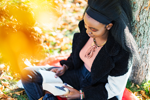 Young adult black woman sitting under maple tree in autumn park and reading book