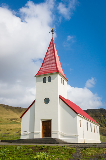 A small wooden white Lutheran church rests on a hillside with jagged mountains behind in  Vik Village, Iceland.