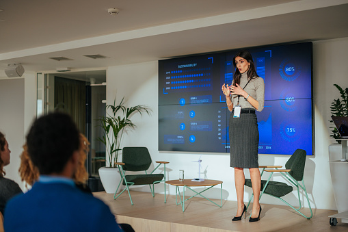 A female team leader is on a stage giving a lecture to her team in the company conference room with the help of charts and graphs on led displays.