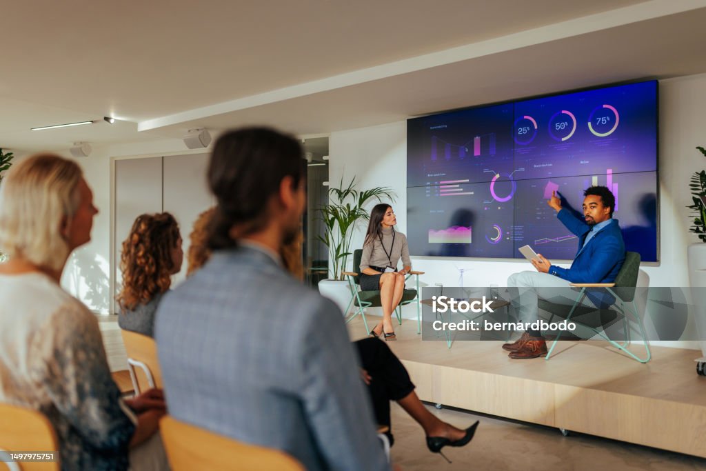 Lecturer presenting sustainable development in conference room. An African American lecturer is presenting a sustainable development strategy to a full auditorium with the help of graphs on a led display. Business Conference Stock Photo Lecturer presenting sustainable development in conference room. An African American lecturer is presenting a sustainable development strategy to a full auditorium with the help of graphs on a led display. Business Conference Stock Photo