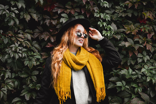 Stylish red haired woman in a black hat, glasses and a yellow scarf stands against a wall of grape leaves. Autumn portrait of a young girl.