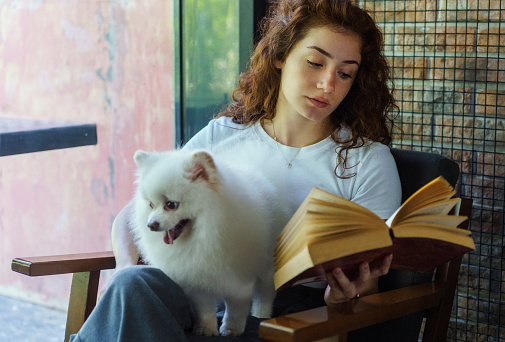 Young woman reading a book with her pomeranian dog in a cafe