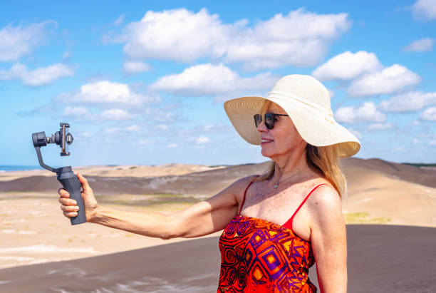 istock Adult mature woman using mobile phone with tripod outdoors on sand dune. 1497982876