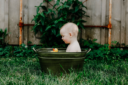 A one~year~old baby boy smiles at his rubber ducky as he sits in a vintage tub in a garden.