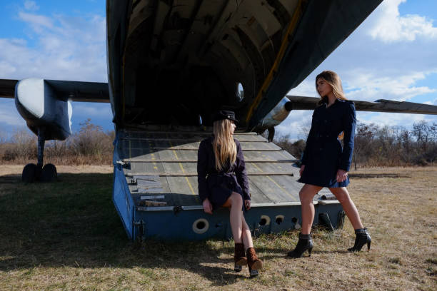 Young blonde elegant woman with hat standing on the stairs of private jet airplane. Bright sunlight is in the background.