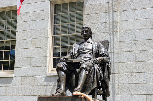 Cambridge, MA, USA - June 29, 2022: A visitor to the Harvard Yard, the oldest part of the Harvard University campus in Cambridge, Massachusetts, touches the foot of the John Harvard statue.