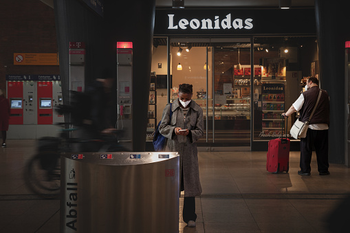 Picture of a young black woman using her smartphone in Koln Hbf train station of Cologne, Germany, while wearing a protective face mask, during the 2020 2021 2022 coronavirus covid 19 health crisis