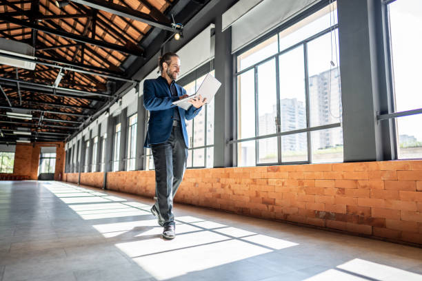 Mature businessman walking and using laptop at empty office for sell at real estate Mature businessman walking and using laptop at empty office for sell at real estate commercial real estate stock pictures, royalty-free photos & images