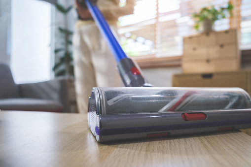 Closeup view of young woman cleaning floor with handheld vacuum cleaner in living room.